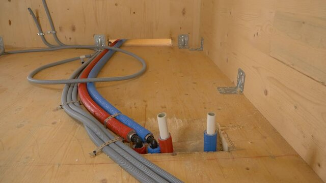 CLOSE UP: Hot and cold water hoses run along wooden floor of a modern wooden house under construction. Corrugated plastic conduits and rubber hoses in utility room of an unfinished housing project.