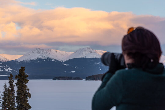 A man with brown hair taking photos of a mountain view scenic spot in northern Canada during winter time March month with snow covered lake, frozen surroundings and mountain peaks. 