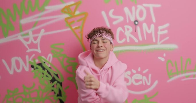 Cool hipster guy dressed in hoodie makes yo gesture moves with rhythm of rap music poses against painted wall draws graffiti being creative street artist. Urban style and young adults concept