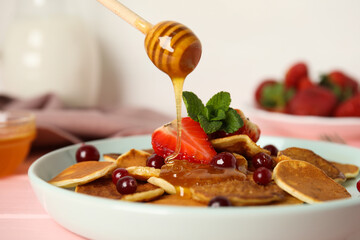 Pouring honey onto cereal pancakes with berries on pink wooden table, closeup