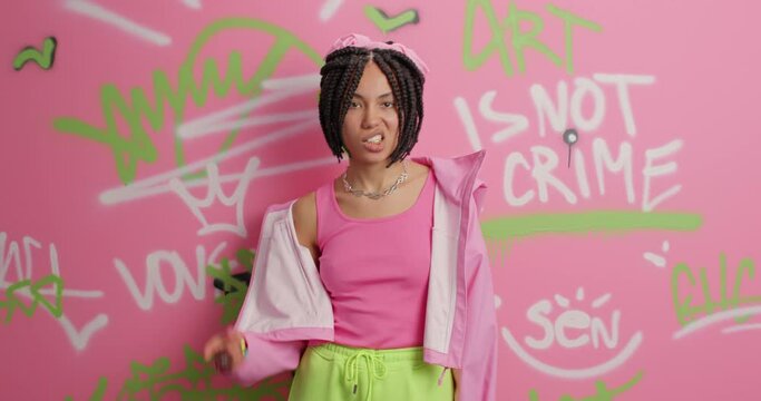 Funky street artist makes yo gesture feels cool enjoys favorite music wears fashionable clothes has dreadlocks poses against colorful graffiti wall. Modern art urban lifestyle millennial generation