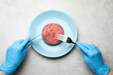 Scientist holding fork and knife over plate with minced cultured meat at light grey lab table, top...