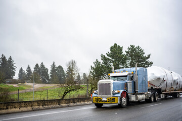 Fototapeta na wymiar Classic blue big rig semi truck with oversize load sign on the bumper transporting cargo on step down semi trailer running on the highway road with wet surface at raining weather