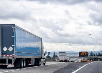 Convoy of big rigs semi trucks with different semi trailers running on the interstate highway with bridge and road sign electronic panel