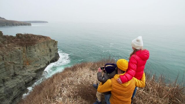 Father is holding his children on the cliff edge and showing them the waves crashing at the rocky coast. The boy is taking pictures with a small camera