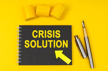 On a yellow background lies a pen, a black notebook with the inscription - CRISIS SOLUTION