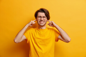 Studio shot of dissatisfied European man clenches teeth cannot focuse because of noise wears spectacles casual t shirt hears heartbreaking scream isolated over yellow background. Unwilling listen