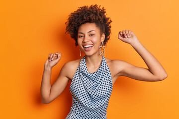 Positive dark skinned curly haired lady dances with arms raised smiles broadly wears fashionable...