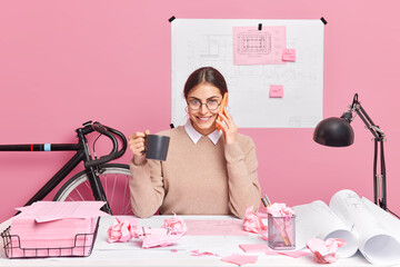 Cheerful office worker works on designing of new office poses in coworking space drinks coffee has telephone conversation isolated over pink background. Skilled female architect prepares project