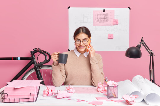 Cheerful office worker works on designing of new office poses in coworking space drinks coffee has telephone conversation isolated over pink background. Skilled female architect prepares project