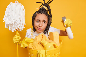 Regular clean up concept. Serious Afro American housewife with braids looks attentively at camera cares about cleanliness holds detergent and mop does housework isolated over yellow background