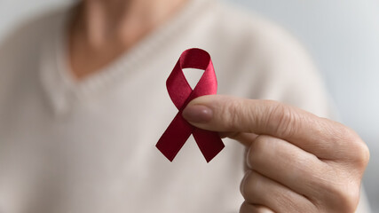 Lady showing hiv or aids awareness symbol, volunteering for charity campaign for prevention immunity disease, cancer, elderly healthcare support. Hand of mature woman holding red ribbon. Close up - obrazy, fototapety, plakaty