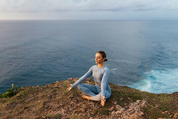 Naklejka na ściany i meble Beautiful woman doing yoga on a cliff, behind an amazing view in the ocean