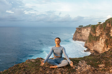 Naklejka na ściany i meble Beautiful woman doing yoga on a cliff, behind an amazing view in the ocean