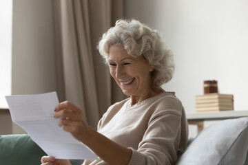 Happy elderly 60s excited about good news, reading paper mail letter in living room. Senior woman...