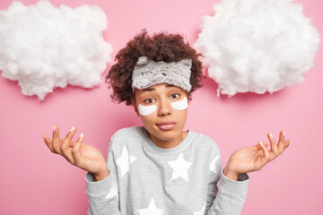 Hesitant curly haired woman spreads palms feels doubt dressed in nightwear applies collagen patches...