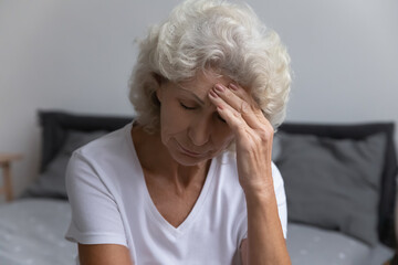 Upset senior lady suffering from headache or high blood pressure, getting bad news, grieving, feeling unwell. Desperate frustrated elderly 60s lady touching head with closed eyes, sitting on bed.