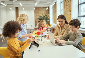 Group of diverse kids working together with young female teacher, using digital devices and playing...