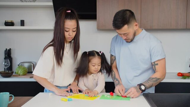 Caring asian parents and adorable baby daughter enjoying playtime together, modeling with colorful plasticine, developing creativity of child through cognitive game while relaxing in domestic kitchen.
