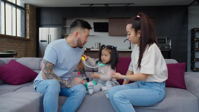 Cheerful adorable asian baby girl playing doctor role game, examining dad's heartbeat and lung with kids stethoscope and having fun during cognitive game while happy family relaxing on sofa at home.