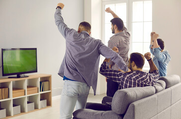 Active adult male friends with arms raised emotionally watching a football match on TV at home....