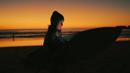 Young woman with surfboard stand at beach during last minutes of sunset, burning bright orange sky, she look at smartphone, device screen is lighting up her face. Millennial use of technology - Powered by Adobe
