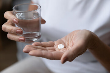 Mature woman holding glass of water and taking painkiller, prescribed antibiotic pill, antidepressant meds, disease symptoms reliever. Hands of woman with white round tablet on palm. Treatment concept