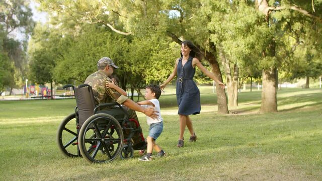 Happy kids and their mom running to disabled retired military dad in park, hugging together. Slow motion. Veteran of war or returning home concept