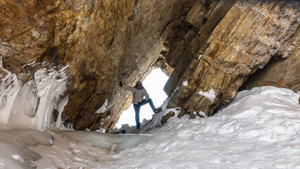 In the opening of the rock, against the background of the sky, there is a man in a protective helmet. The leg is bent, the hand is raised, touches the wall. At the base of the cliff - ice, snow