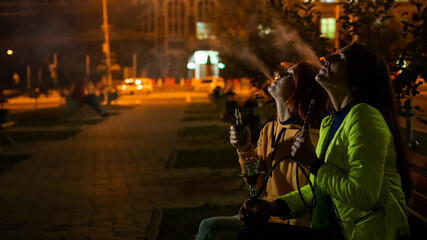Obraz premium Two caucasian women smoke vape and hookah outdoors at night.
