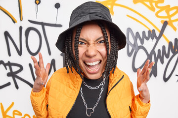 Urban casual style. Cheeky woman with dreadlocks raises hands exclaims angrily wears fashionable clothes poses near graffiti wall at street belongs to hooligan gang. Youth and street concept - obrazy, fototapety, plakaty