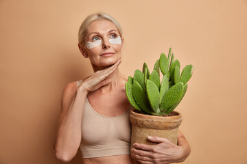 Portrait of thoughtful middle aged European woman touches jawline gently applies collagen patches under eyes holds potted cactus wears top isolated over brown background. Women age skin care concept