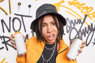 Discontent teenage girl with braids holds aerosol cans for making graffiti dressed in yellow jacket black hat metal chains around neck enjoys urban street liifestyle smirks face. Funky hipster - obrazy, fototapety, plakaty