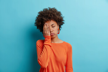 Fototapeta na wymiar Bored sleepy woman with curly hair makes face palm feels tired or exhausted stands alone closes eyes wears casual orange jumper isolated over blue wall. Attractive female model has gloomy expression