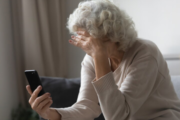Sad senior lady using mobile phone at home, sitting on couch, covering face with hand. Frustrated...
