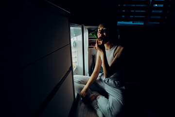 Portrait of a beautiful girl taking food from the refrigerator at night. Night food, overeating concept. Diet, dieting. Beauty young woman eating dessert in refrigerator in night kitchen.