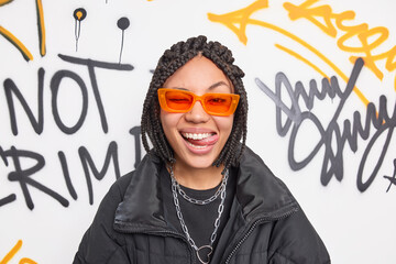 Cheerful fancy teenage girl wears trendy orange suglasses and black jacket sticks out tongue has dreadlocks hairstyle expresses positive emotions poses against graffiti wall. Swag and style concept - obrazy, fototapety, plakaty