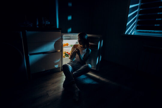 a hungry girl sits with her elbows on an open refrigerator holding a bottle of milk in her hands. The image was shot in a low key to convey night time.