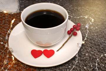 A white coffee cup stands on a saucer with two red hearts among the lights. Warm sweater and top