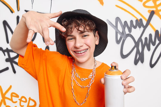 Happy boy teenagers gestures cool sign smiles positively wears braces on teeth wears fashionable clothes holds aerosol spray poses against graffiti wall. Urban clothing style. Street art concept