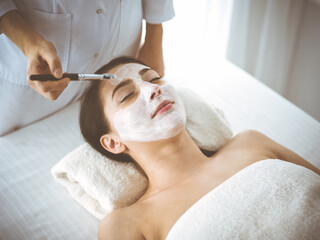 Beautiful brunette woman enjoying applying cosmetic mask with closed eyes comfortable and blissful. Relaxing treatment in medicine and spa center concepts