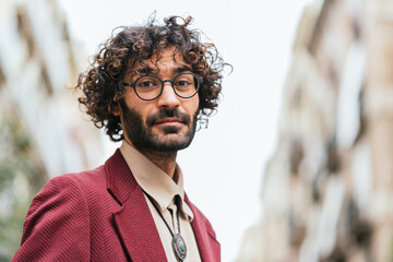 horizontal portrait of young caucasian man on the street looking at camera. Wear trendy casual clothes and glasses. He has curly hair and a beard