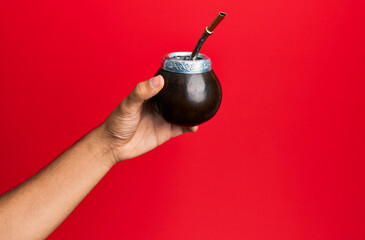 Hand of hispanic man holding cup of mate infusion beverage over isolated red background.