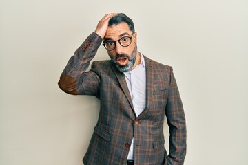 Fototapeta premium Middle age man with beard and grey hair wearing business jacket and glasses surprised with hand on head for mistake, remember error. forgot, bad memory concept.