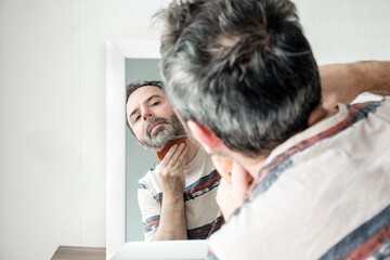 Mature man cutting his own grey beard and moustache at home with scissors and comb during coronavirus isolation.