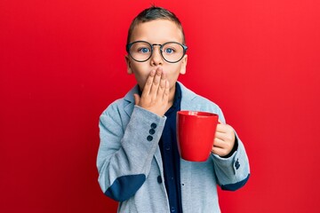 Little kid boy wearing glasses drinking from a red mug covering mouth with hand, shocked and afraid...