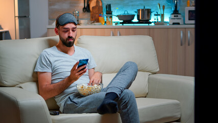 Bored man sitting on couch holding popcorn bowl while texting message on social network using...