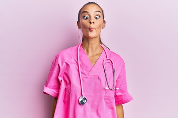 Young hispanic woman wearing doctor uniform and stethoscope making fish face with lips, crazy and comical gesture. funny expression.