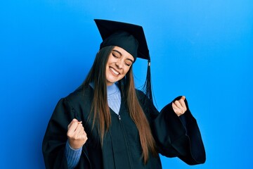 Beautiful brunette young woman wearing graduation cap and ceremony robe very happy and excited...