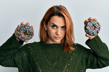Hispanic young woman holding tasty colorful doughnuts skeptic and nervous, frowning upset because...